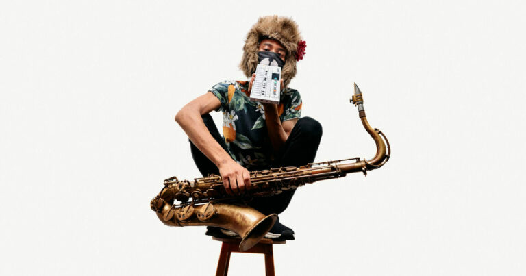 Tomoki Sanders seated on a stool holding a saxophone, wearing a floral shirt and a furry hat with flower. Their face is obscured by a small electronic device and an animal-themed mask.