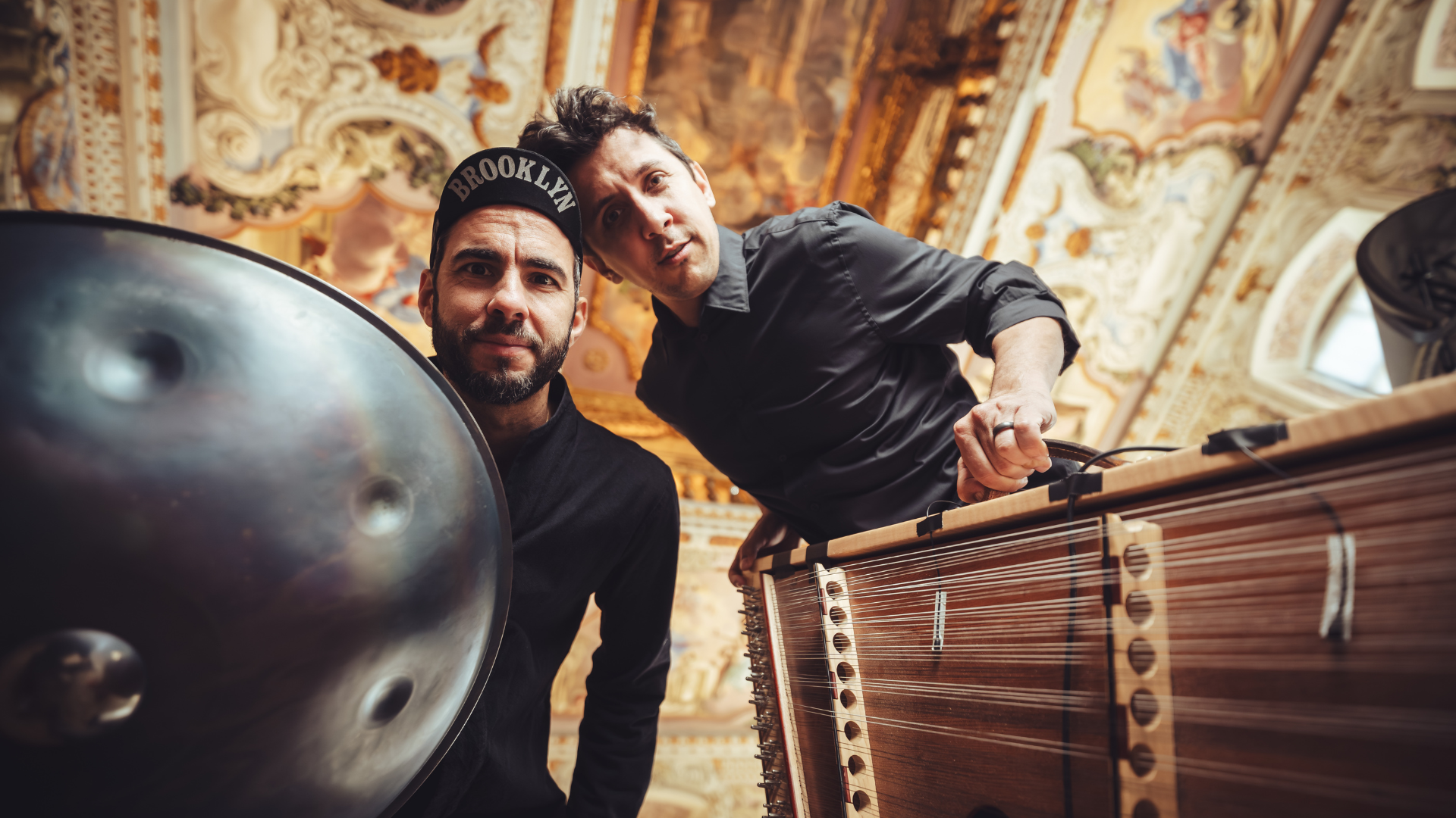 Manu Delago ja Maz XT poseeraavat koristeellisessa salissa. Etualalla näkyvät handpan-rumpu ja dulcimer, joita muusikot soittavat.