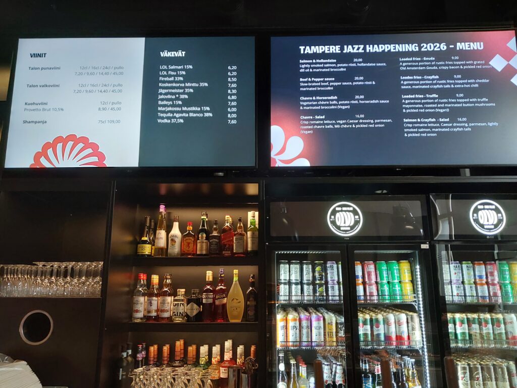 Klubin baaritiski, jonka yläpuolella digitaaliset juoma- ja ruokalistat. Hyllyillä alkoholijuomia ja kylmäkaapeissa tölkki- ja pullotuotteita.