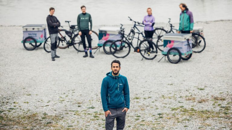 Manu Delago etualalla, taustalla ryhmä polkupyörineen ja tavarapyöräkärryineen järven rannalla.